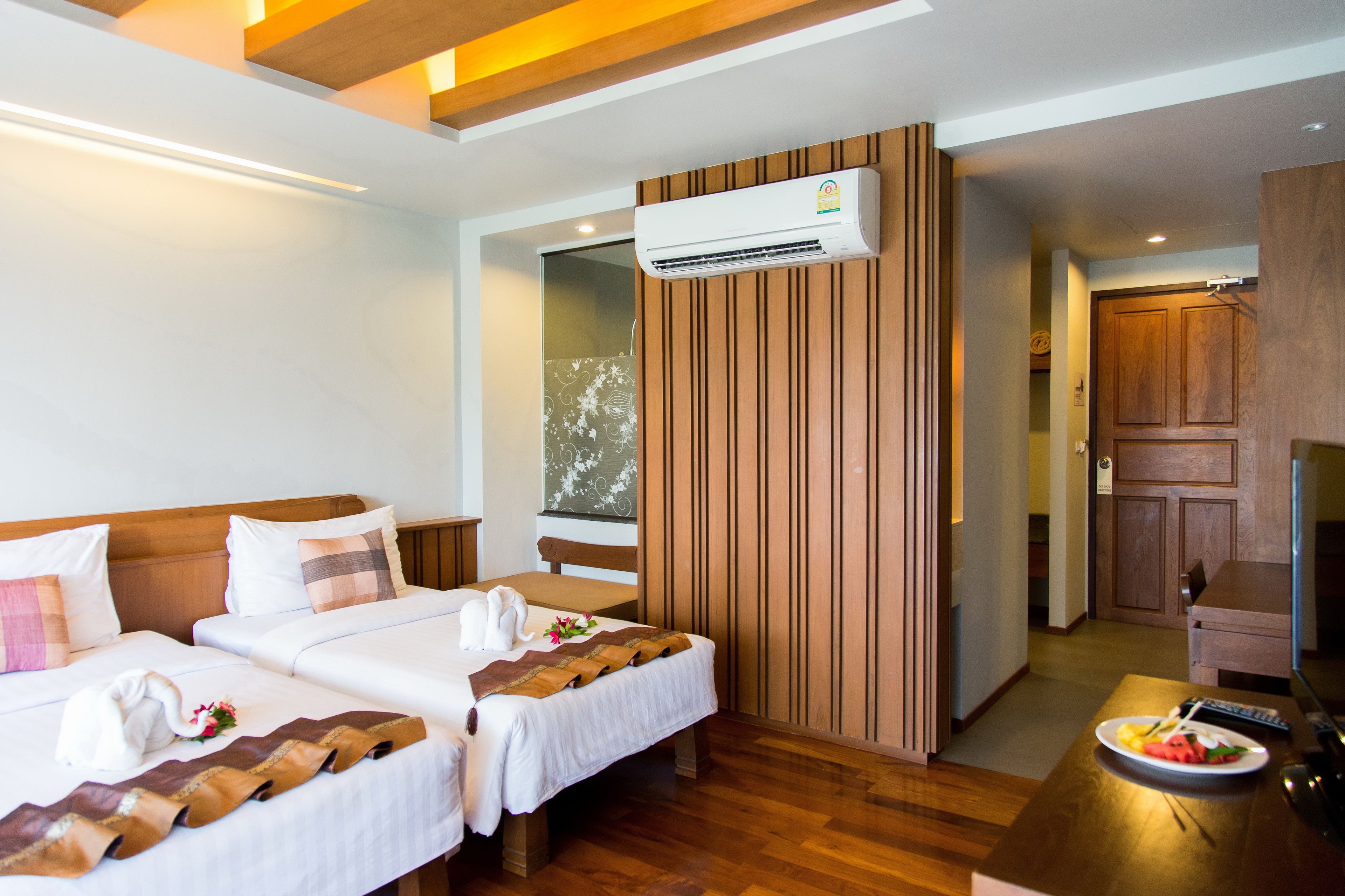 Habitación de hotel con dos camas, un separador de madera, un equipo de aire acondicionado, una mesa de comedor con un plato de comida y un televisor.