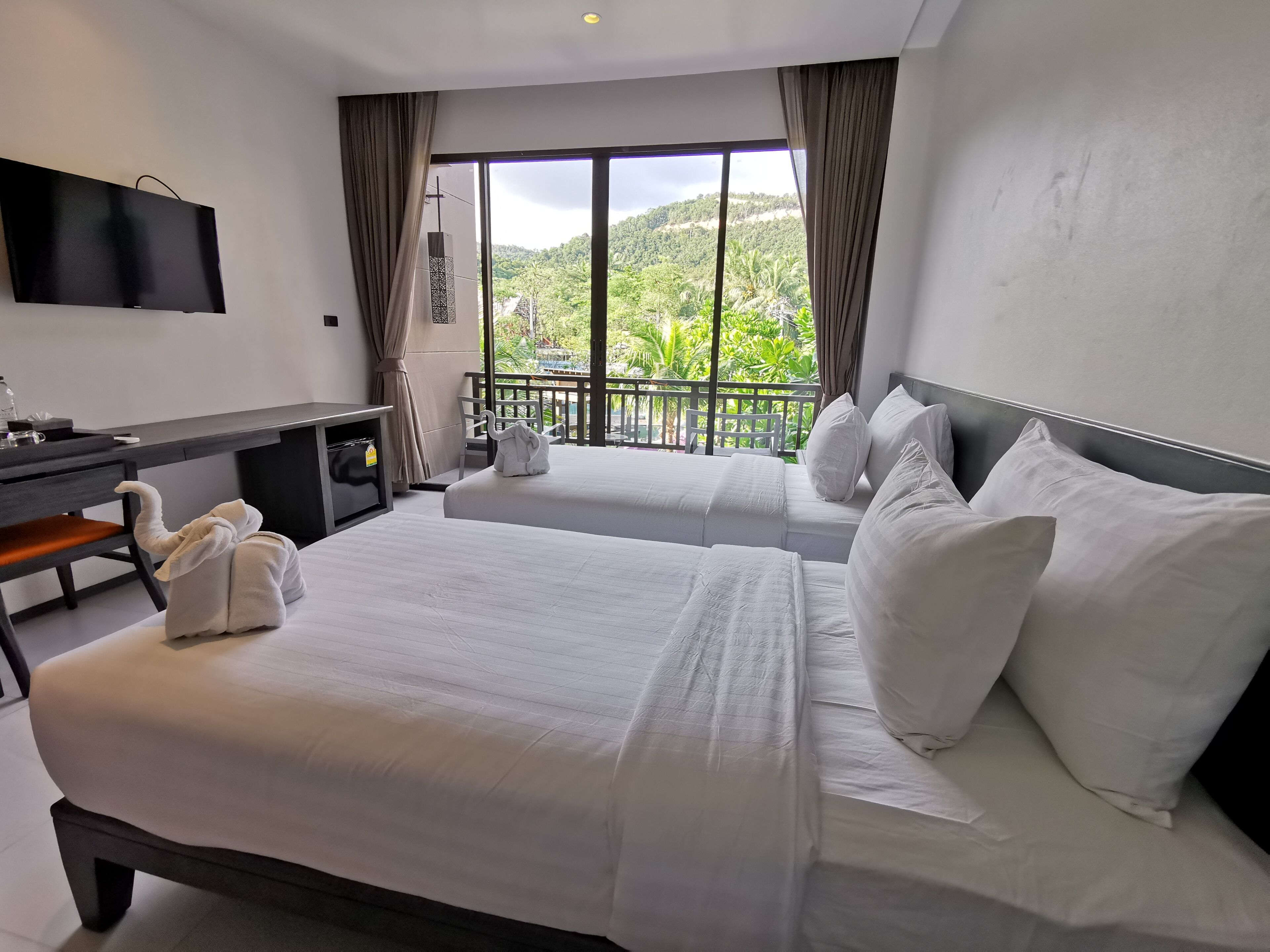 Ein Hotelzimmer mit zwei Betten, einem Schreibtisch, einem Fernseher und einem Balkon mit Blick auf Grünflächen.