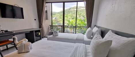 A hotel room with two beds, a desk, a TV, and a balcony with a view of greenery.