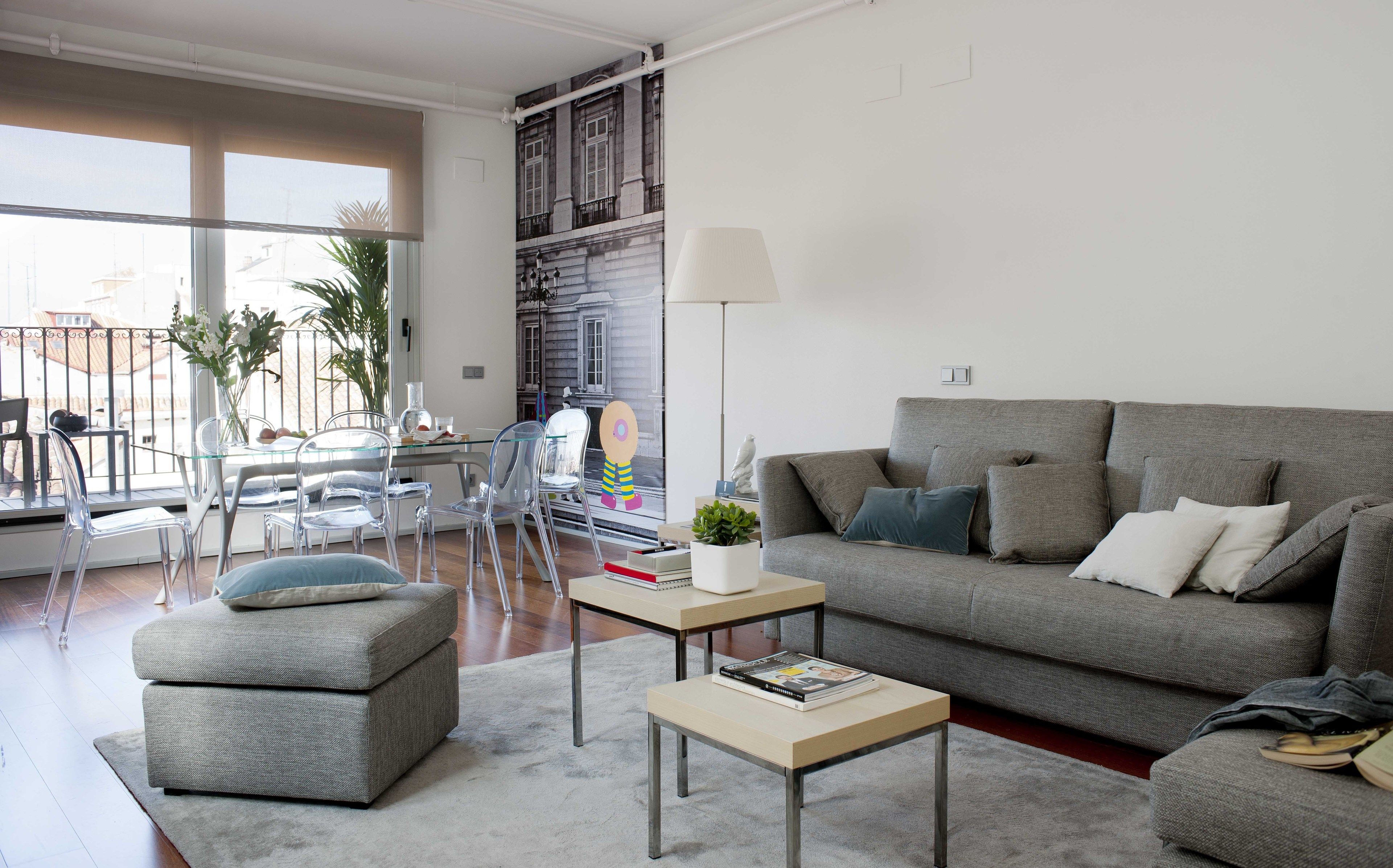 Una sala de estar moderna con un sofá gris, sillas transparentes, una mesa de vidrio y un ventanal con vistas a edificios.