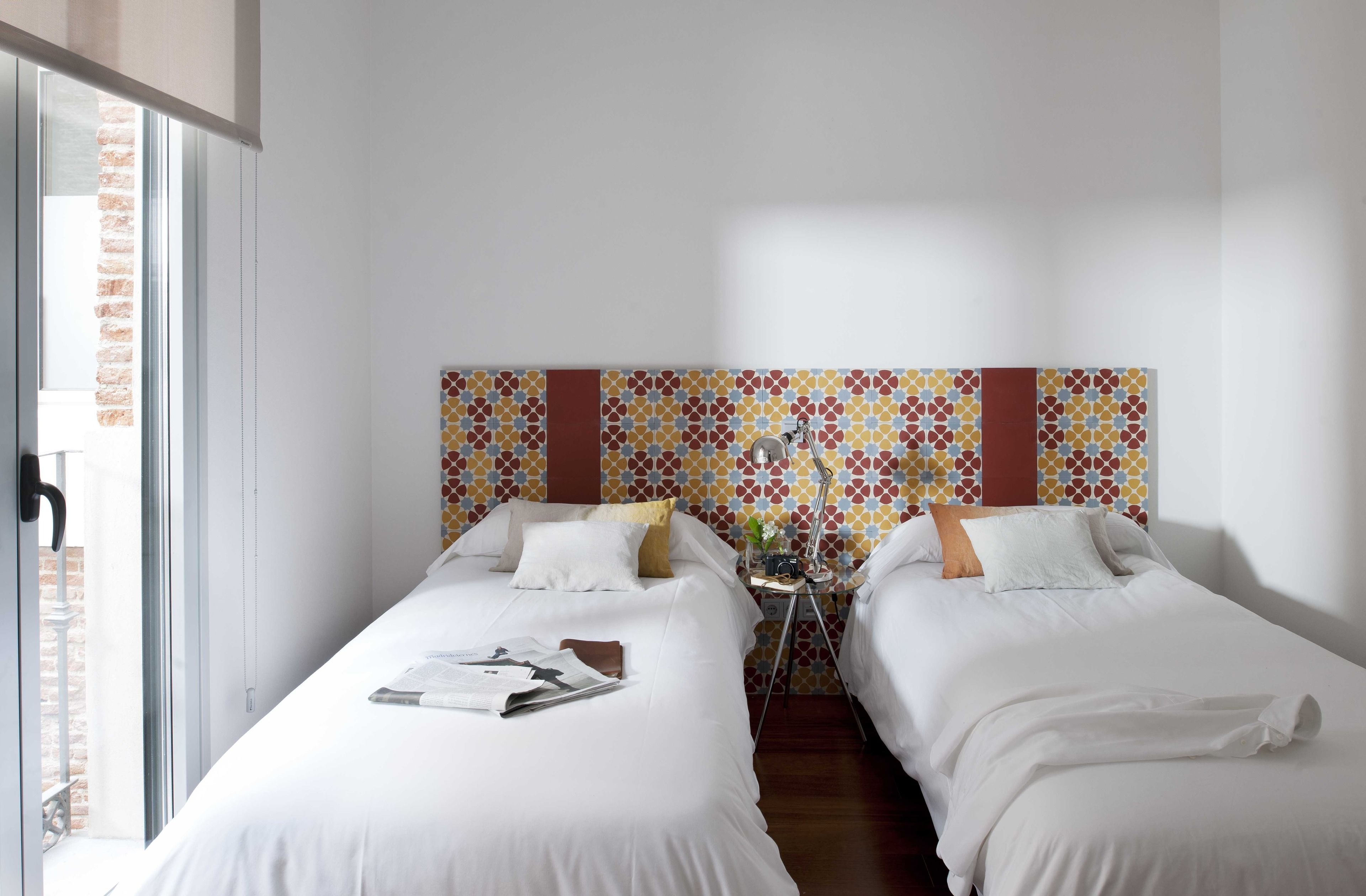 Habitación de hotel con dos camas, un cabecero estampado, una mesita con una planta y una ventana con vista a un muro de ladrillo.