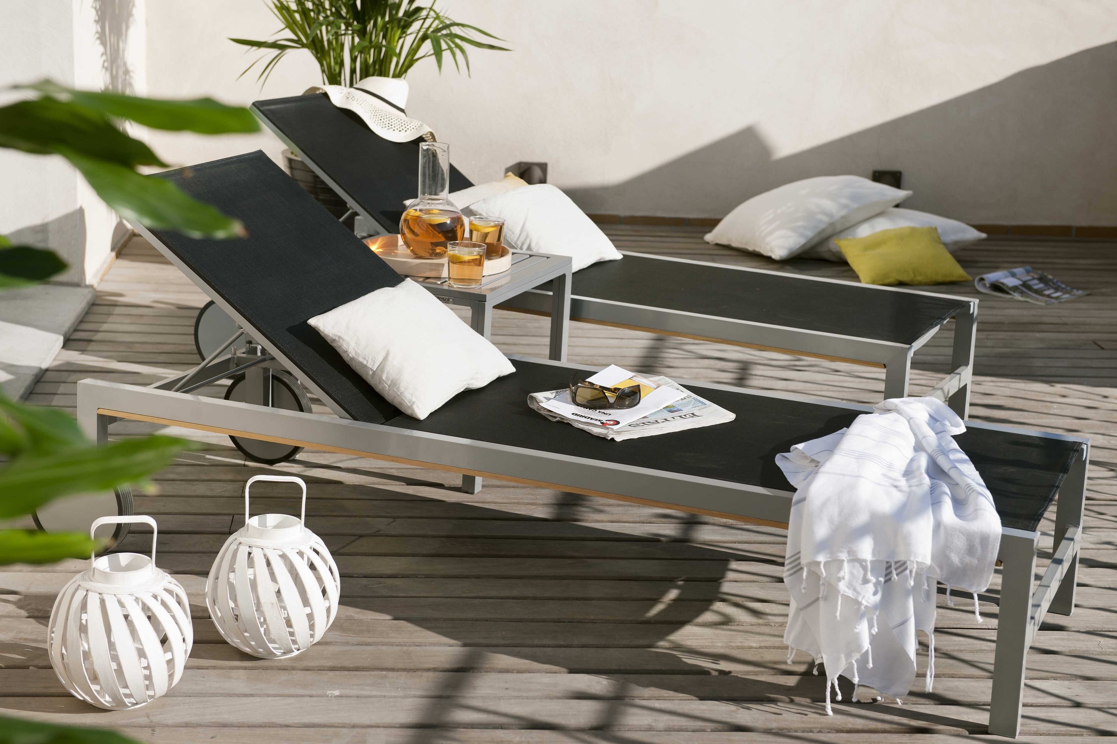 Un área de descanso al aire libre iluminada por el sol, con dos sillones reclinables, una mesa con bebidas y faroles decorativos.