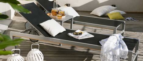 A sunlit outdoor lounge area with two reclining chairs, a table with drinks, and decorative lanterns.