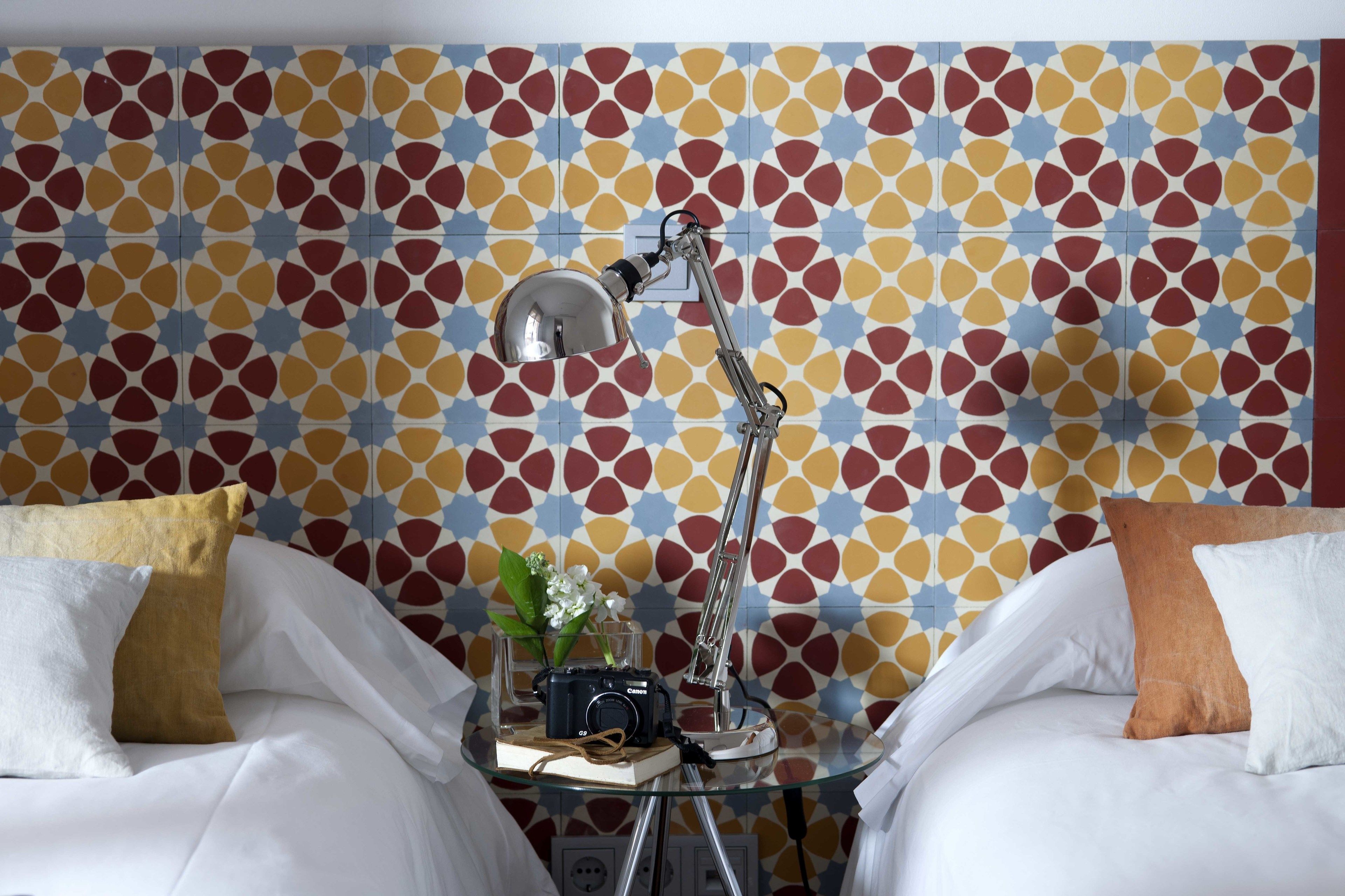 Una habitación de hotel con pared estampada, dos camas con ropa blanca, una mesa de vidrio con una cámara, una lámpara y un jarrón con flores.