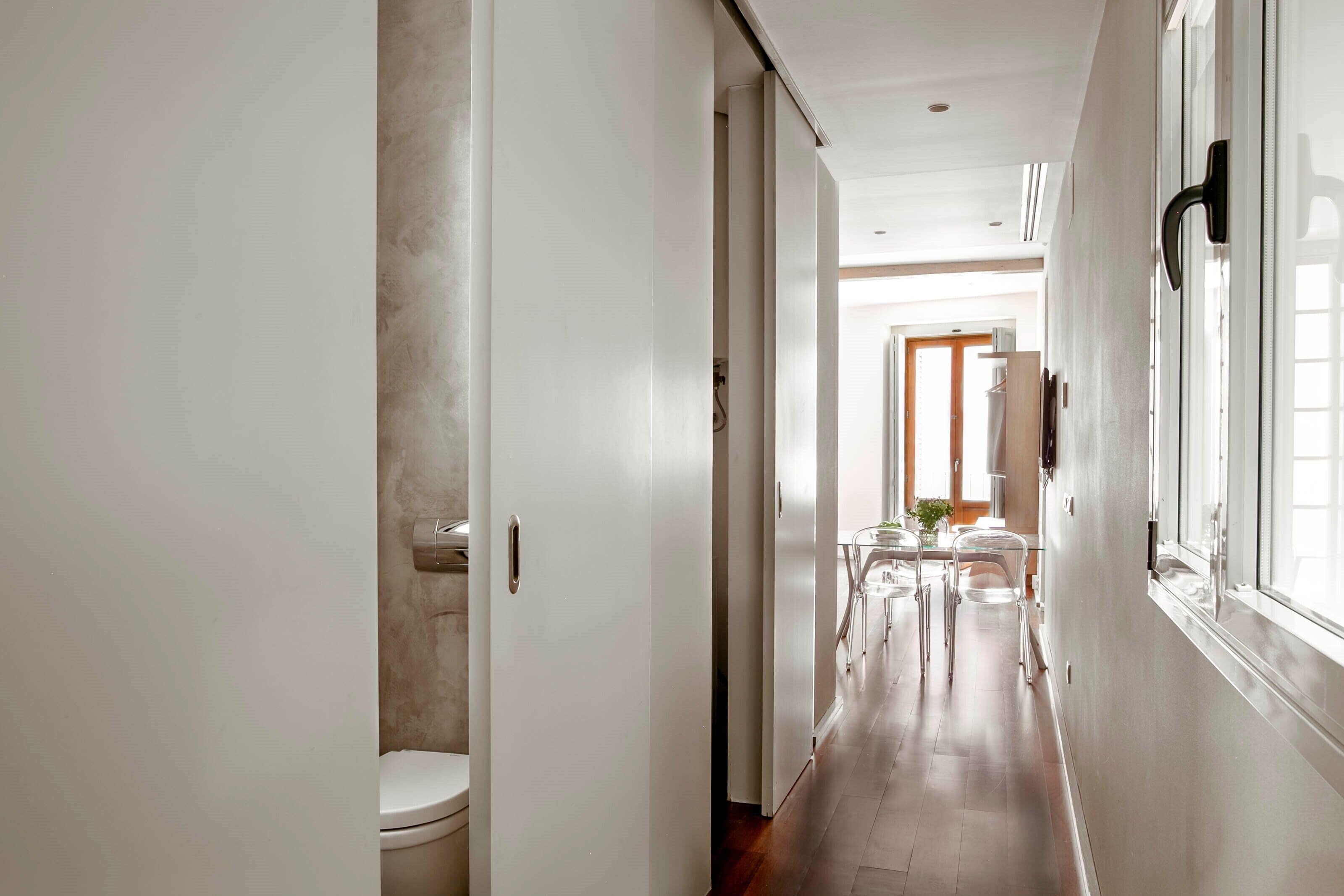 A narrow hallway with a toilet on the left, a dining area with chairs and a table in the center, and a window on the right.