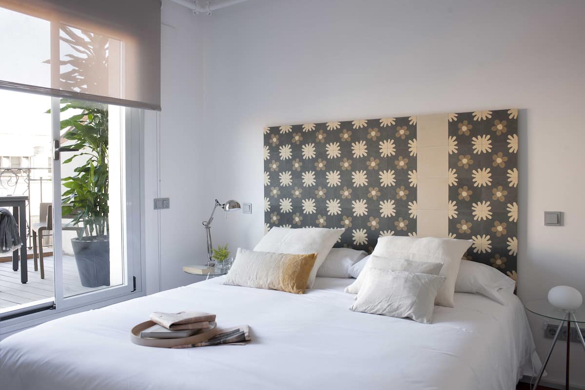 A bedroom with a large bed, white bedding, a patterned headboard, a bedside table with a lamp, and a view of an outdoor area with a plant and furniture.