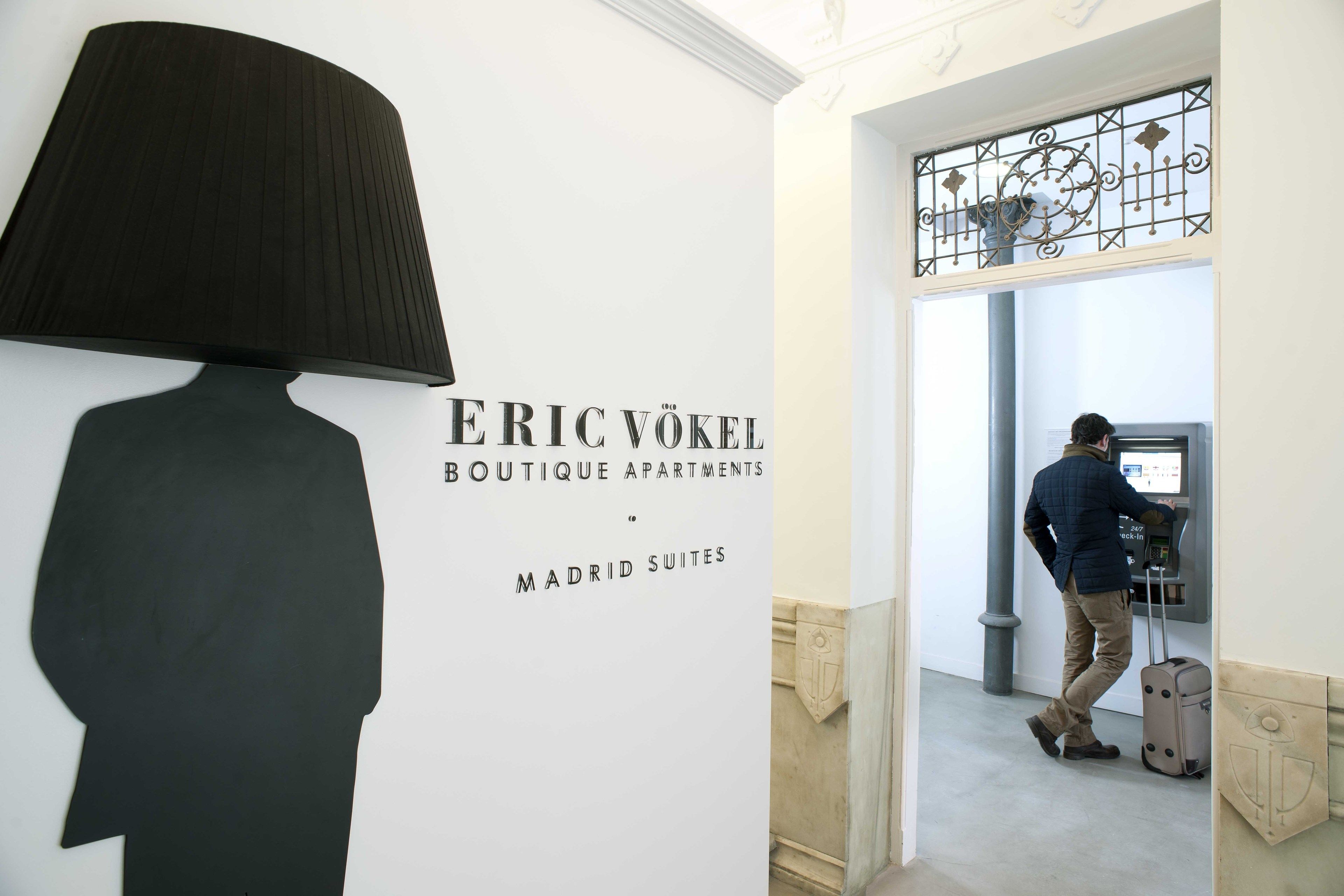 A hotel interior with a wall-mounted lamp, a man at a self-service kiosk, and a sign for Eric Vokel Boutique Apartments in Madrid Suites.