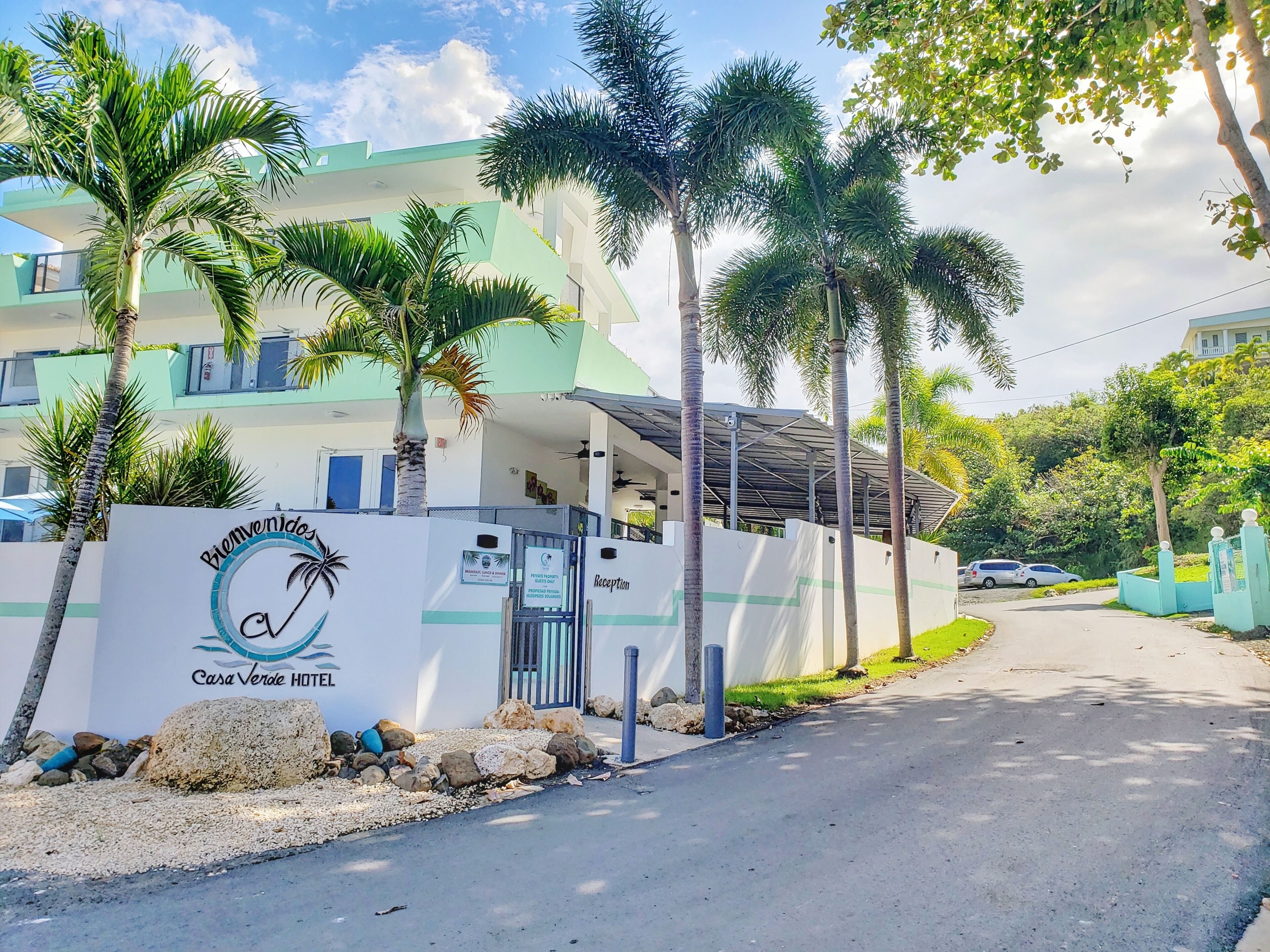 A hotel entrance with a reception desk, palm trees, and a sign reading "Casa Verde Hotel".