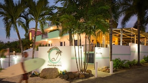 A hotel entrance with a sign for Casa Verde Hotel, palm trees, and a person carrying a surfboard. - Casa Verde Hotel - Steps to the Beach | Pool, Restaurant & Bar (Rincon)