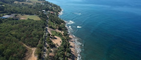 An aerial view of a coastal area with a beach, dense vegetation, and a clear view of the ocean.