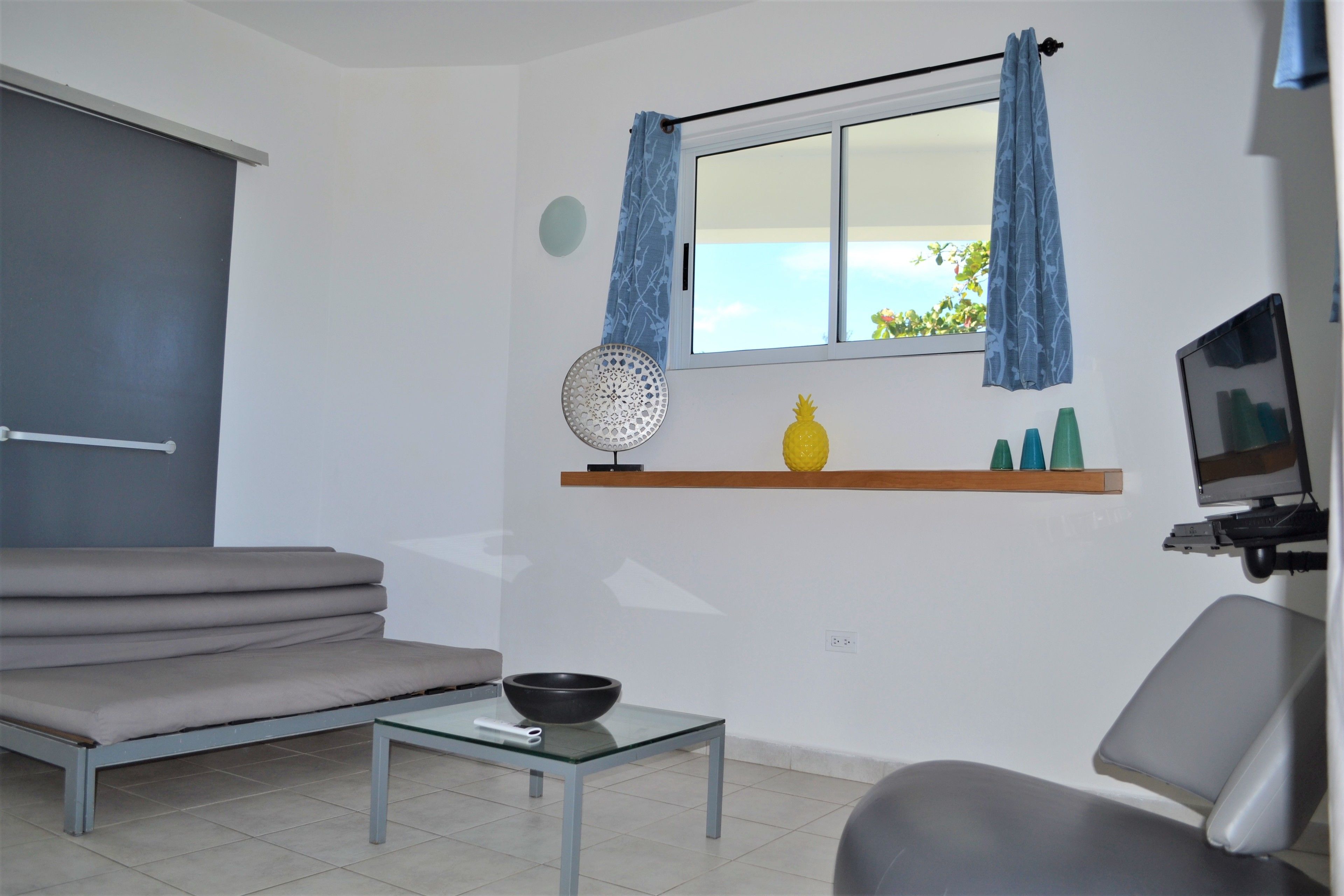 Un salón moderno con un sofá, una mesa de vidrio, una mesita con un cuenco, una ventana con cortinas y un televisor fijado a la pared.