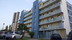 A multi-story residential building with balconies, a parking area, and a street lamp.