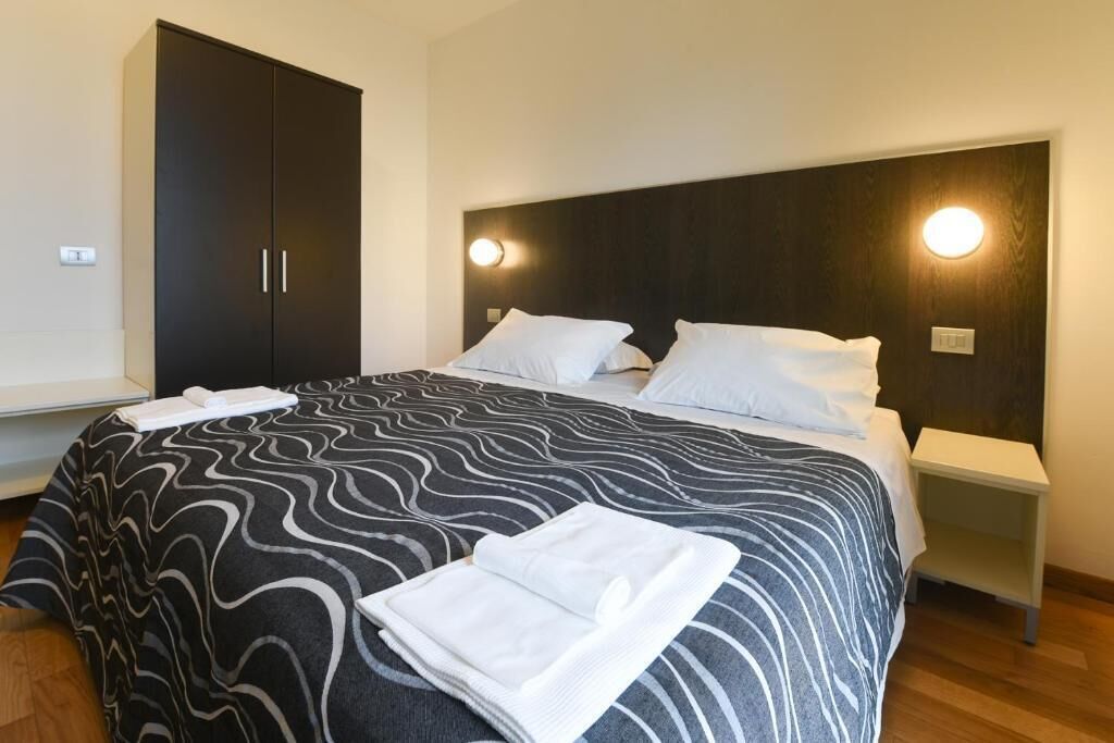 A hotel room with a large bed, a dark wardrobe, bedside tables, and a headboard with wall-mounted lights.