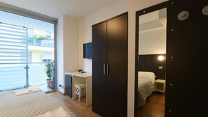 A bedroom with a bed, a desk, and a wardrobe. There is a window with blinds, a potted plant, and a mirror.