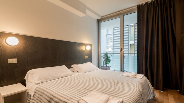 A hotel room with a large bed, a bedside table, a nightstand, a window with curtains, and a view of buildings outside.