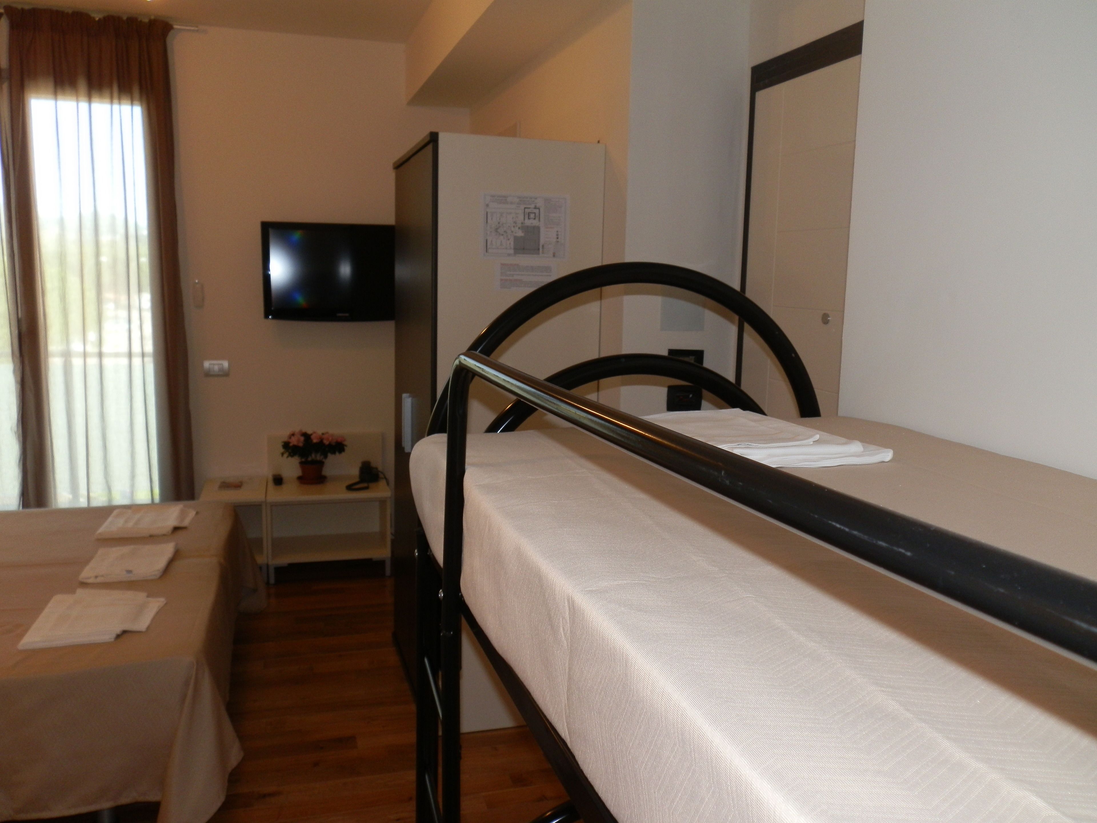 A single bed with a metal frame, a bedside table with a lamp, a flat-screen TV mounted on the wall, and a window with curtains.