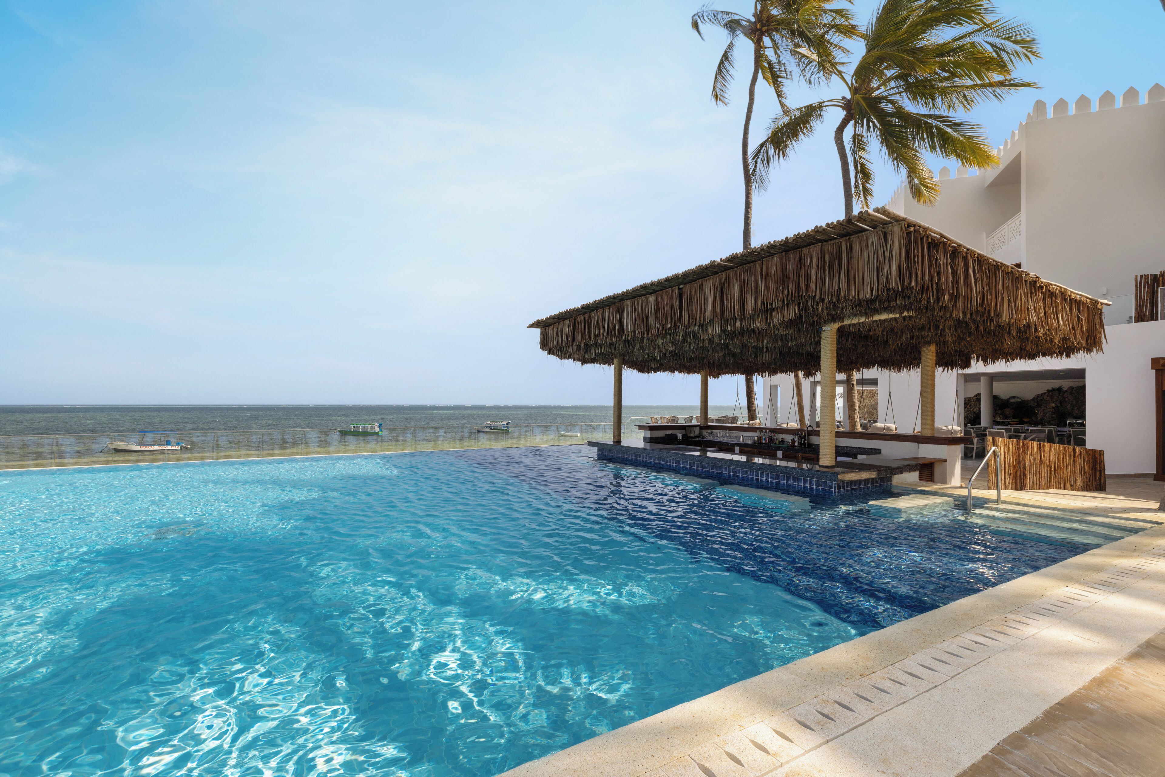 A clear blue pool with a thatched roof structure, palm trees, and a view of the ocean.