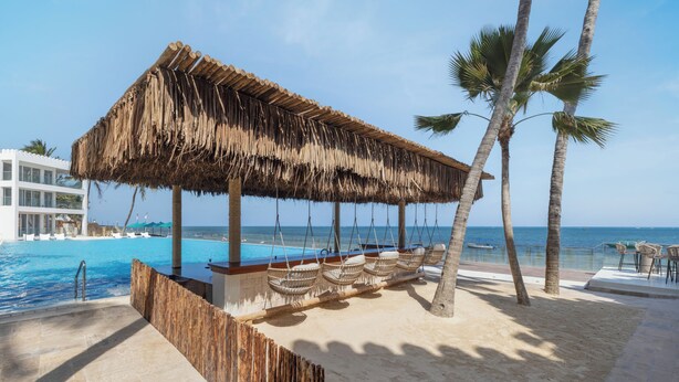 A beachside pool area with a thatched roof structure, palm trees, and a view of the ocean. - Bamburi Beach Hotel - All Inclusive (Mombasa)