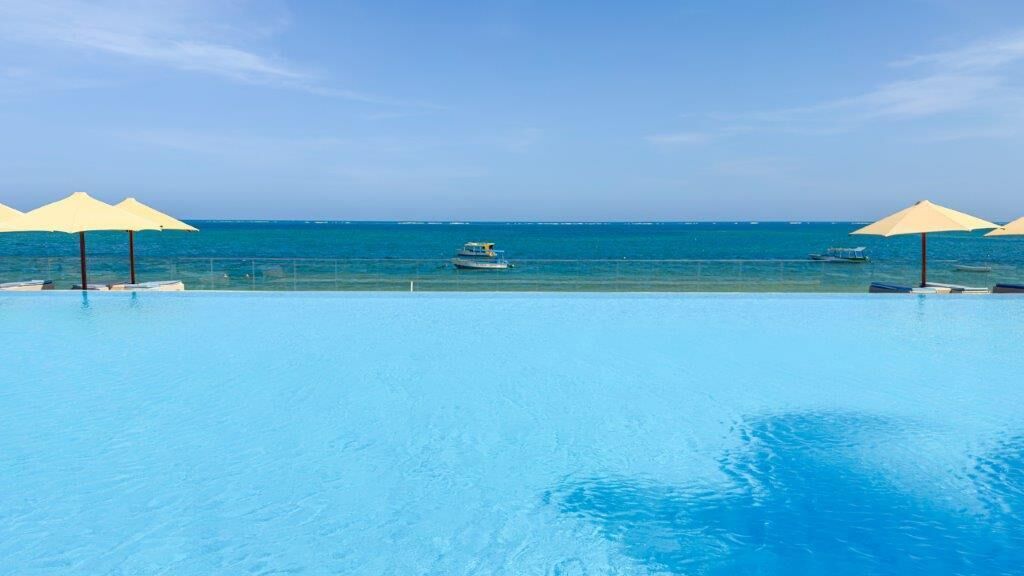 A clear blue pool with umbrellas, a boat in the background, and a view of the sea.