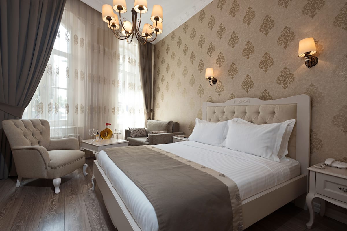 A hotel room with a large bed, a sitting area with a chair and a table, a chandelier, and patterned wallpaper.
