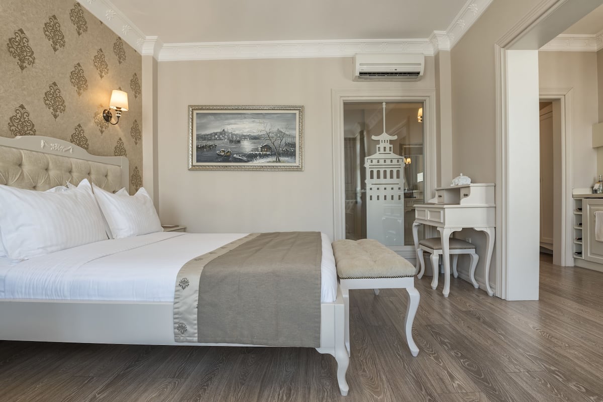 A bedroom with a large bed, a desk, and a chair. There is a painting on the wall and an air conditioning unit above the desk.
