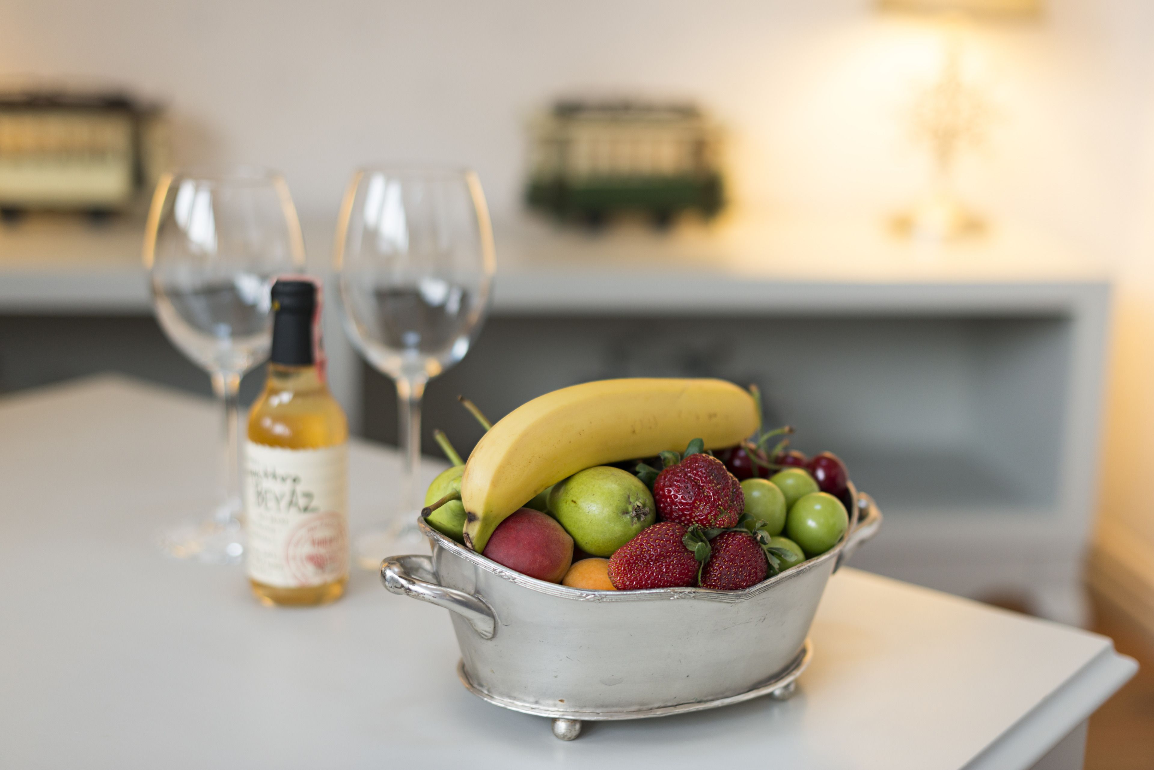 A metal bowl filled with fruits and a bottle of wine on a table.