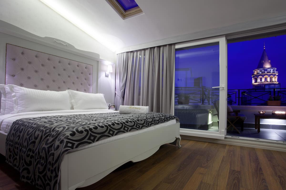 A bedroom with a large bed, a headboard, a nightstand, and a view of a balcony with a railing and a building in the background.