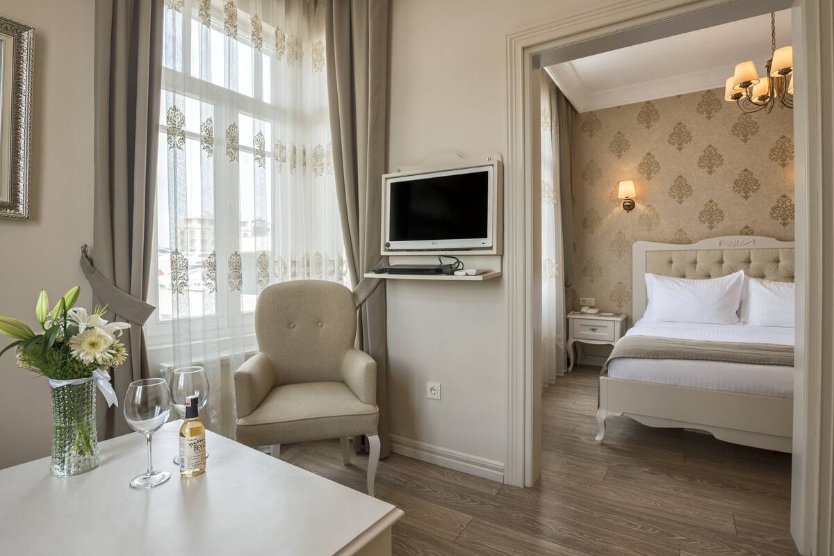 A hotel room with a bed, a TV mounted on the wall, a chair, a table with a vase of flowers and wine glasses, and a window with curtains.