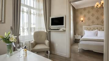 Une chambre d’hôtel avec un lit, un téléviseur fixé au mur, une chaise, une table avec un vase de fleurs et des verres à vin, et une fenêtre avec des rideaux.