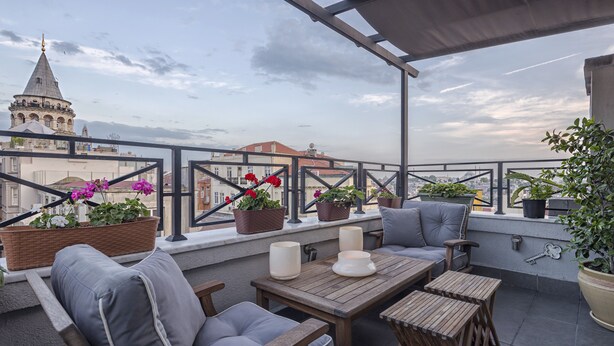 A rooftop terrace with a wooden table, two grey cushioned chairs, potted plants, and a view of a cityscape including a church tower. - Louis Appartements Galata (Istanbul)