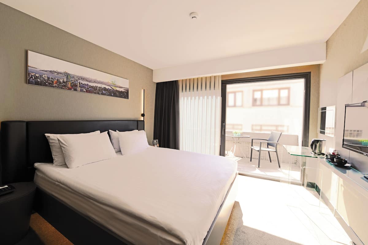 A hotel room with a large bed, a desk with a chair, a television, and a balcony with a view.