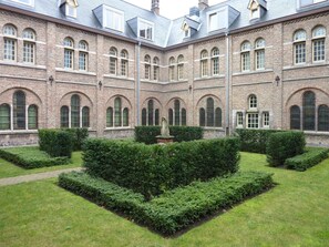 Ein Innenhof mit einer zentralen Fontäne, umgeben von ordentlich geschnittenen Hecken und einem Backsteinhaus mit Bogenfenstern.