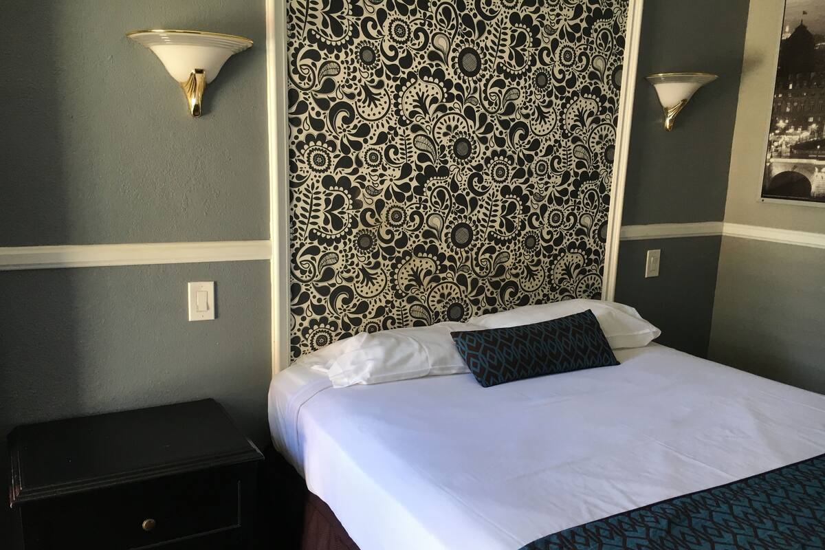 A hotel room with a patterned headboard, a bed with white linens and a blue patterned throw, and a nightstand with drawers.