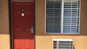 Uma porta vermelha com placa de identificação, uma janela com persianas fechadas e um ar condicionado branco numa parede bege.