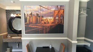 Een hotelkamer met een groot stadsgezicht schilderij, een ronde spiegel, een wastafel, een klein tafeltje met stoelen en een compacte koelkast.