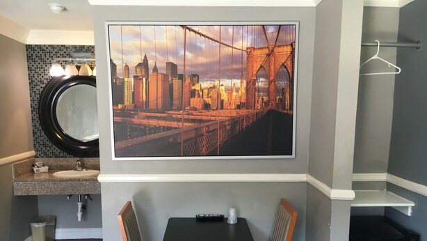Quarto de hotel com uma grande pintura da paisagem urbana, um espelho redondo, uma pia, uma pequena mesa com cadeiras e um refrigerador compacto.