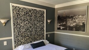 Quarto de hotel com cabeceira estampada, cama com roupa de cama azul e branca, mesa de cabeceira e quadro com vista para a cidade.