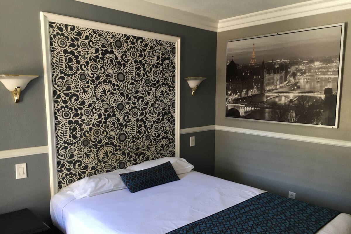 A hotel room with a patterned headboard, a bed with white and blue bedding, a nightstand, and a framed picture of a cityscape.