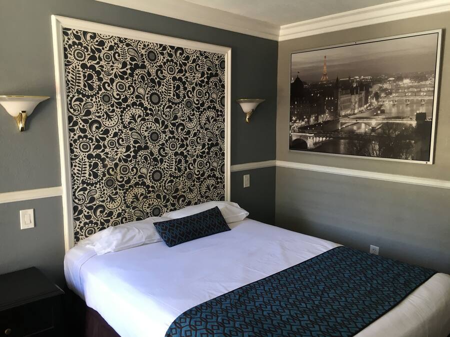 A hotel room with a patterned headboard, a bed with white and blue bedding, a nightstand, and a framed picture of a cityscape.