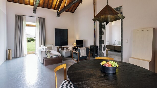 Una sala moderna con una mesa de comedor, un sofá y un televisor. Hay una ventana con cortinas, techo de madera y una lámpara colgante.