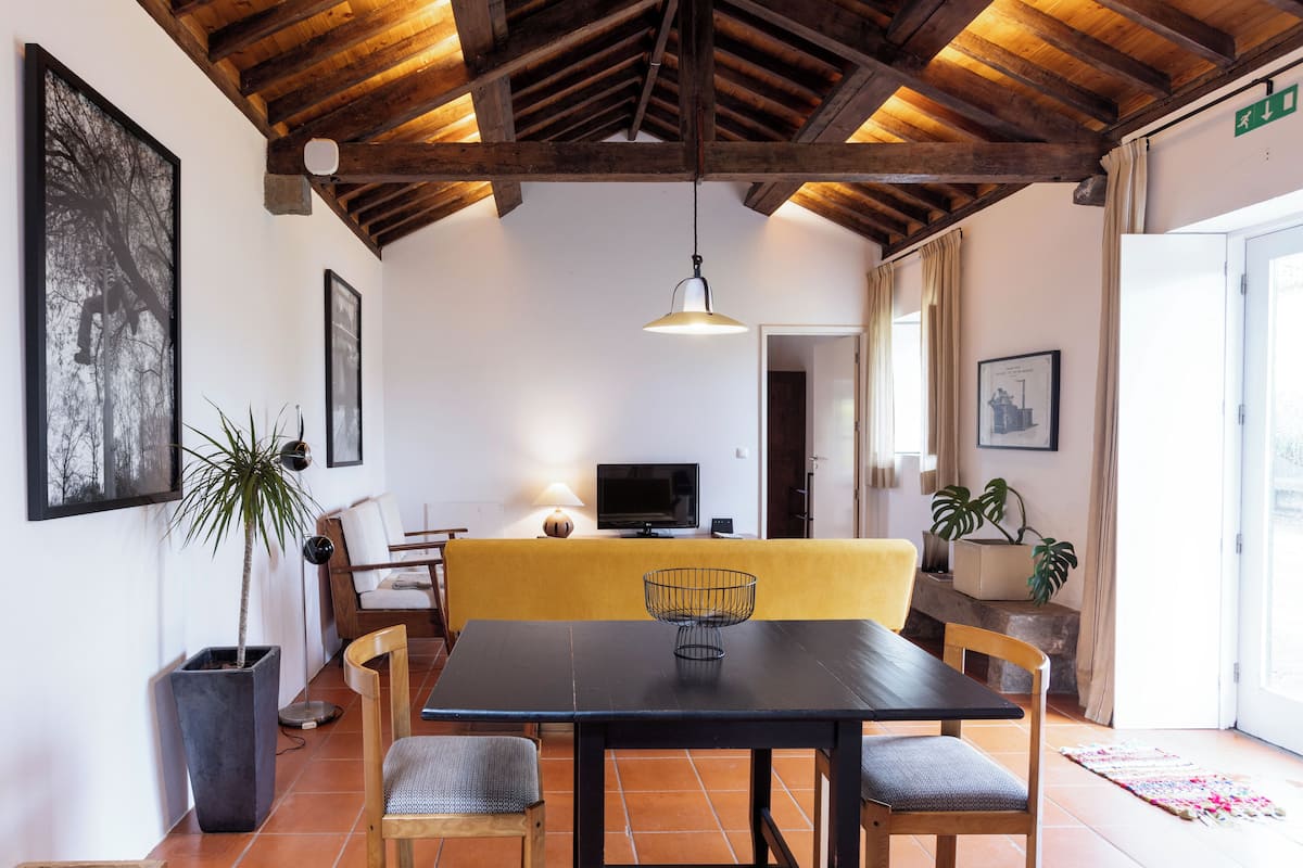 A dining area with a black table, four chairs, a yellow bench, a TV, and a ceiling with exposed beams.