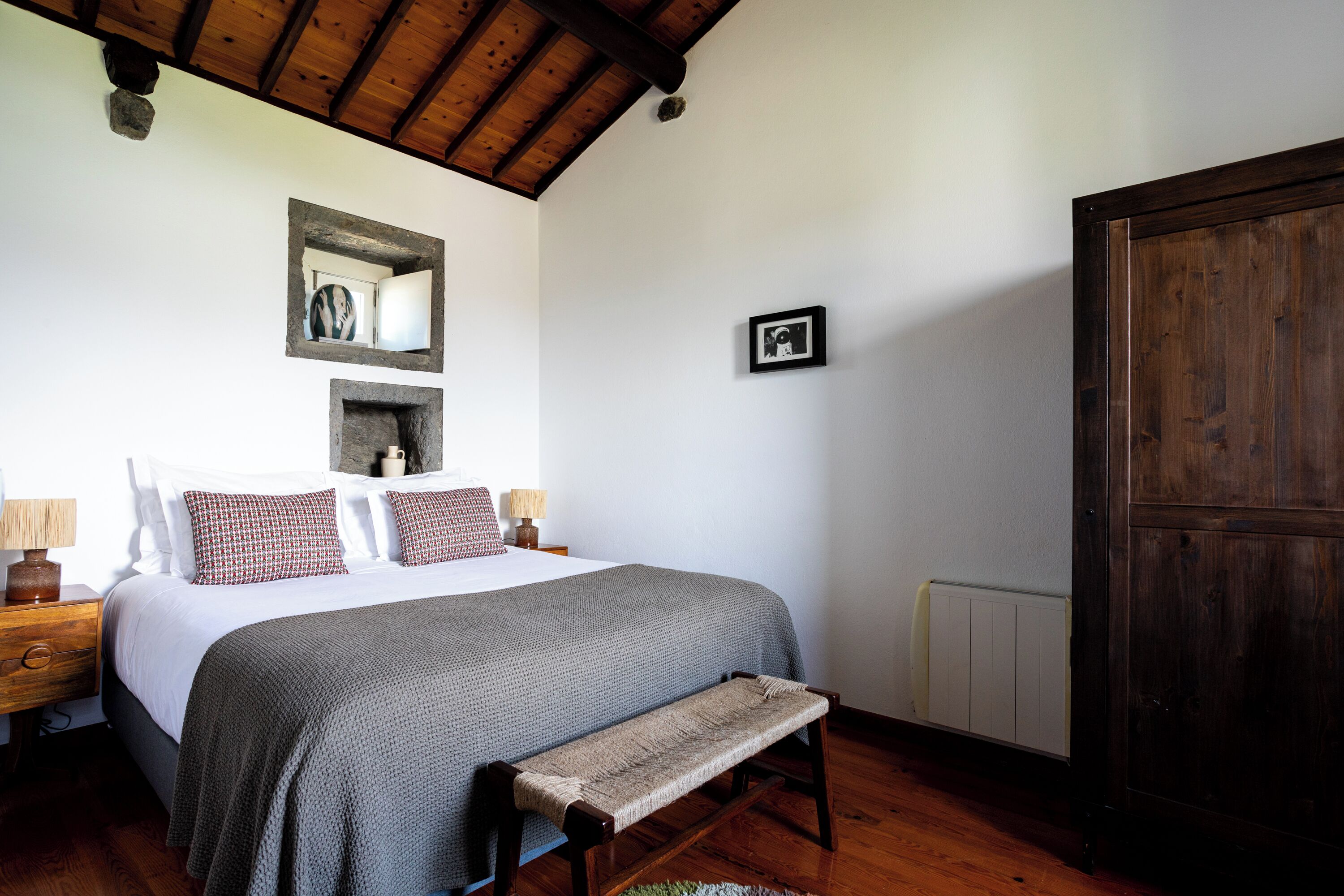 A bedroom with a wooden ceiling, a bed with a grey blanket, a bench, a nightstand with a lamp, and a wardrobe.