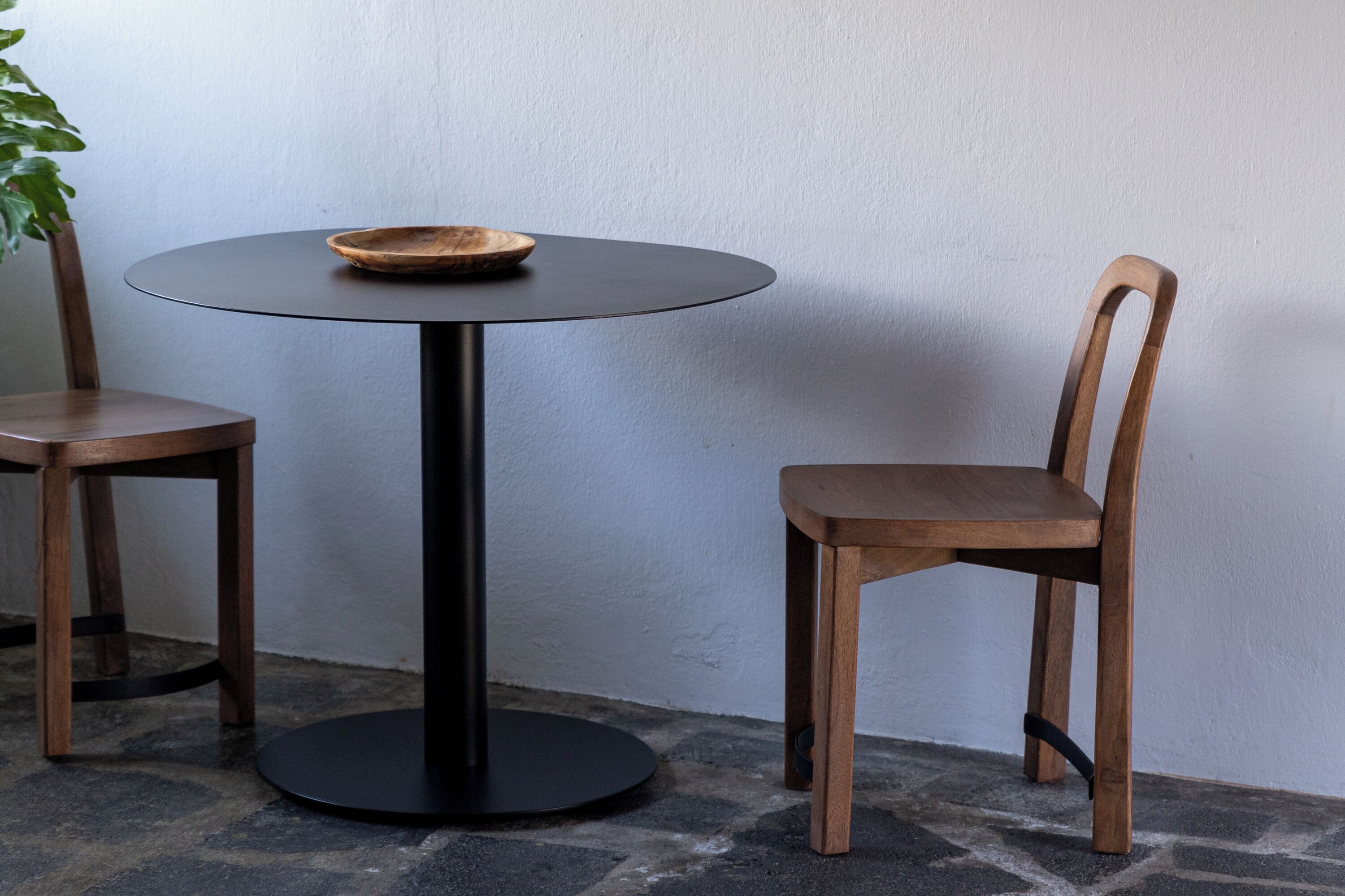 Ein runder Tisch mit schwarzem Fuß und einer Holzschüssel auf der Tischplatte, daneben zwei Stühle mit Metallbeinen.