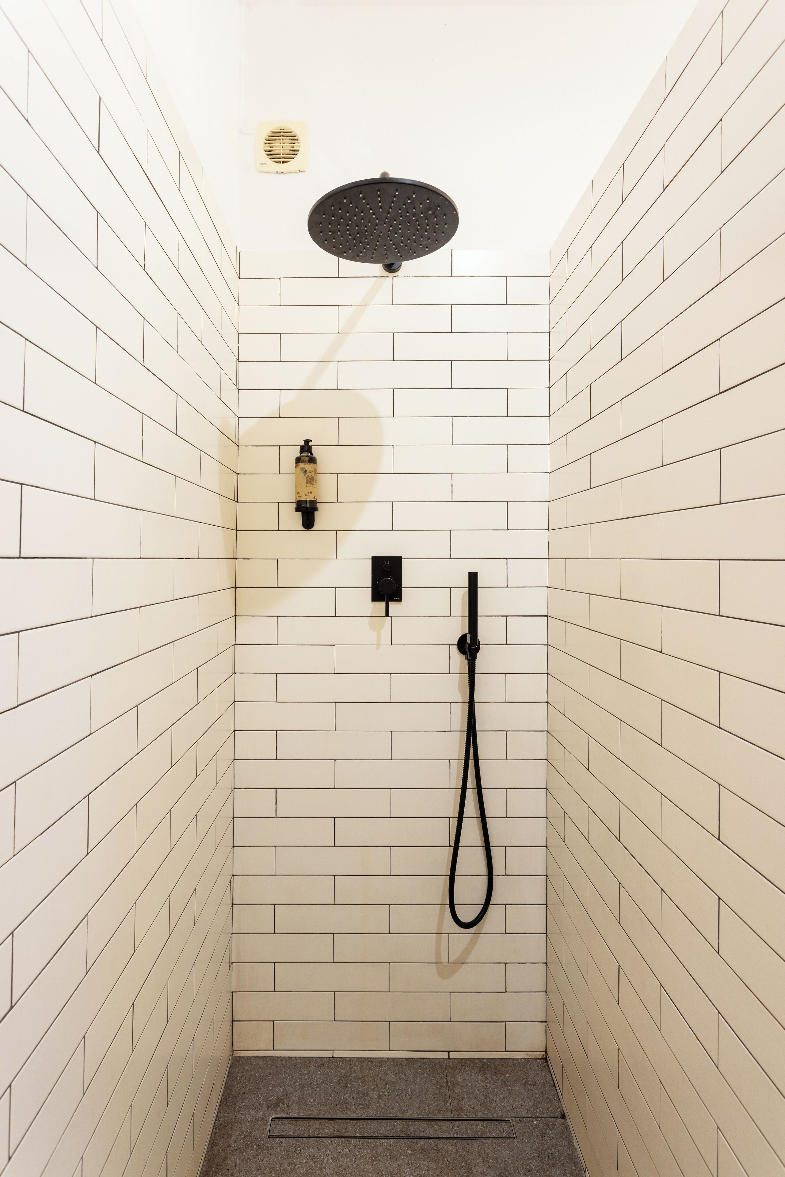 A small, white-tiled shower stall with a handheld showerhead and a handheld shower hose.
