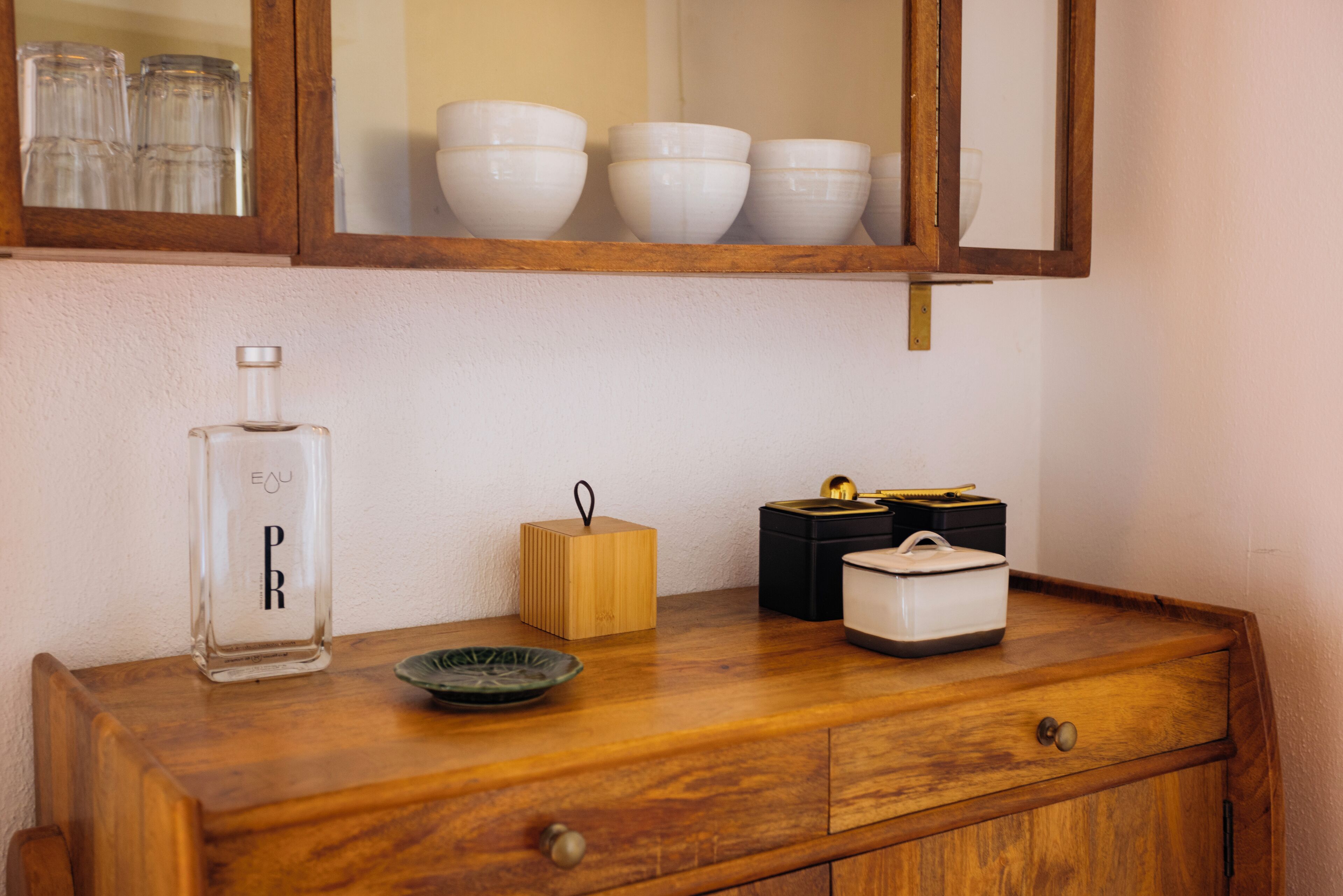 Een houten dressoir met een spiegel erboven, een fles met de tekst 'Eau PR', een klein houten doosje en twee zwart-wit containers.