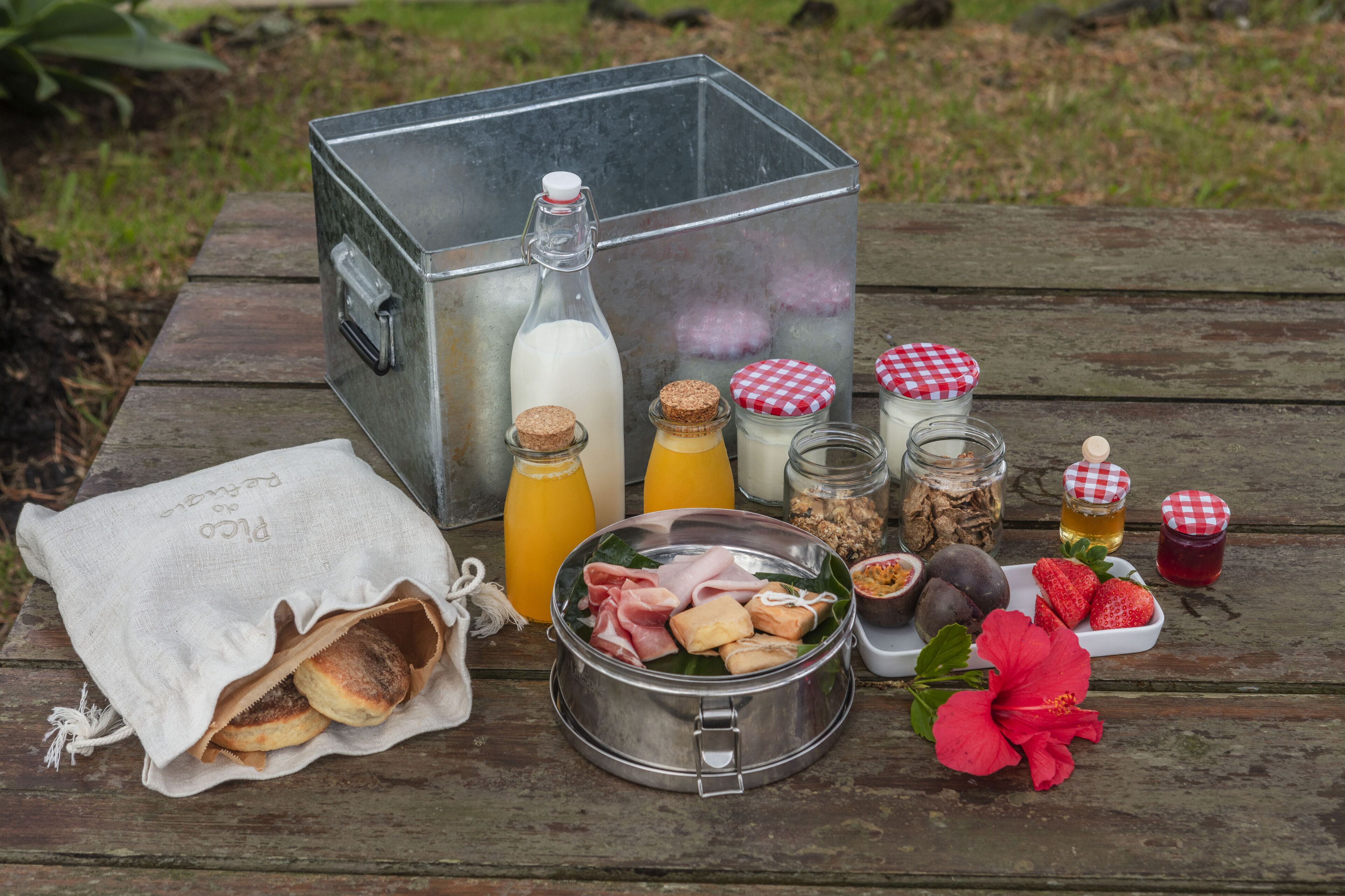 Une table en bois est dressée pour un pique-nique, avec un récipient en métal, des bouteilles, des bocaux, un sac en tissu et divers aliments.