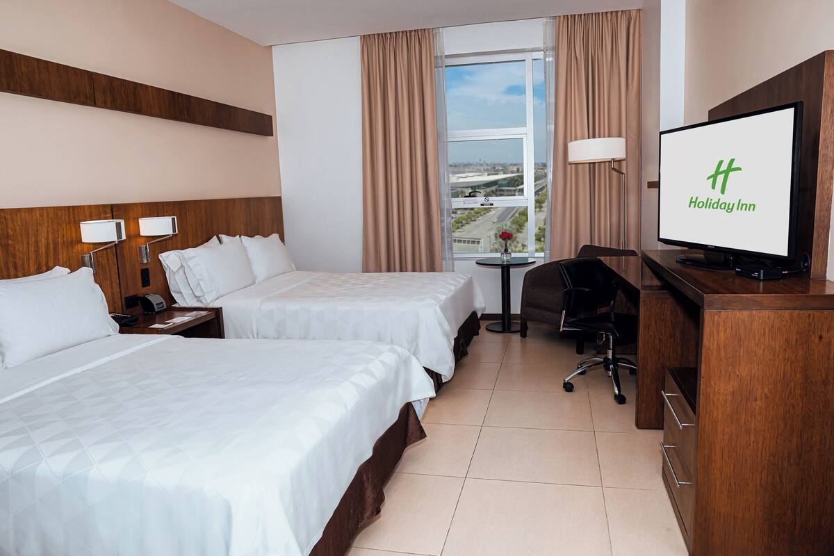 Een hotelkamer met twee bedden, een bureau met een computer, een televisie waarop het Holiday Inn logo staat en een raam met uitzicht op de stad.