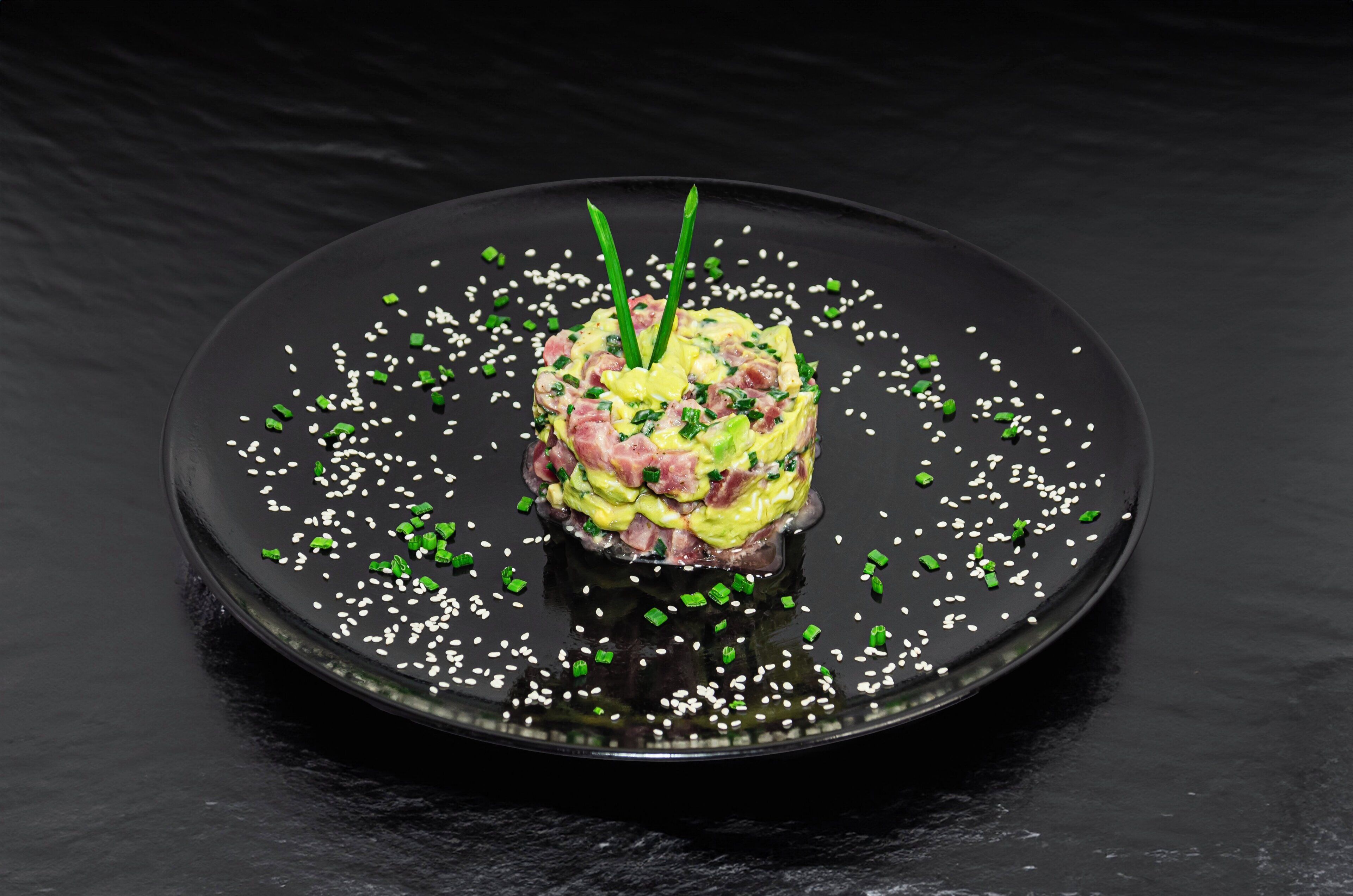 Un plat servi dans une assiette noire, garni de graines de sésame blanches et d’herbes vertes.