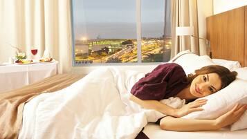 A hotel room with a bed, a nightstand with a glass of red wine, and a view of a cityscape at night.