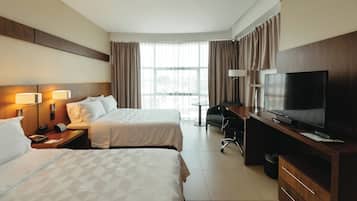 A hotel room with two beds, a desk with a TV, a chair, a lamp, and a window with curtains.